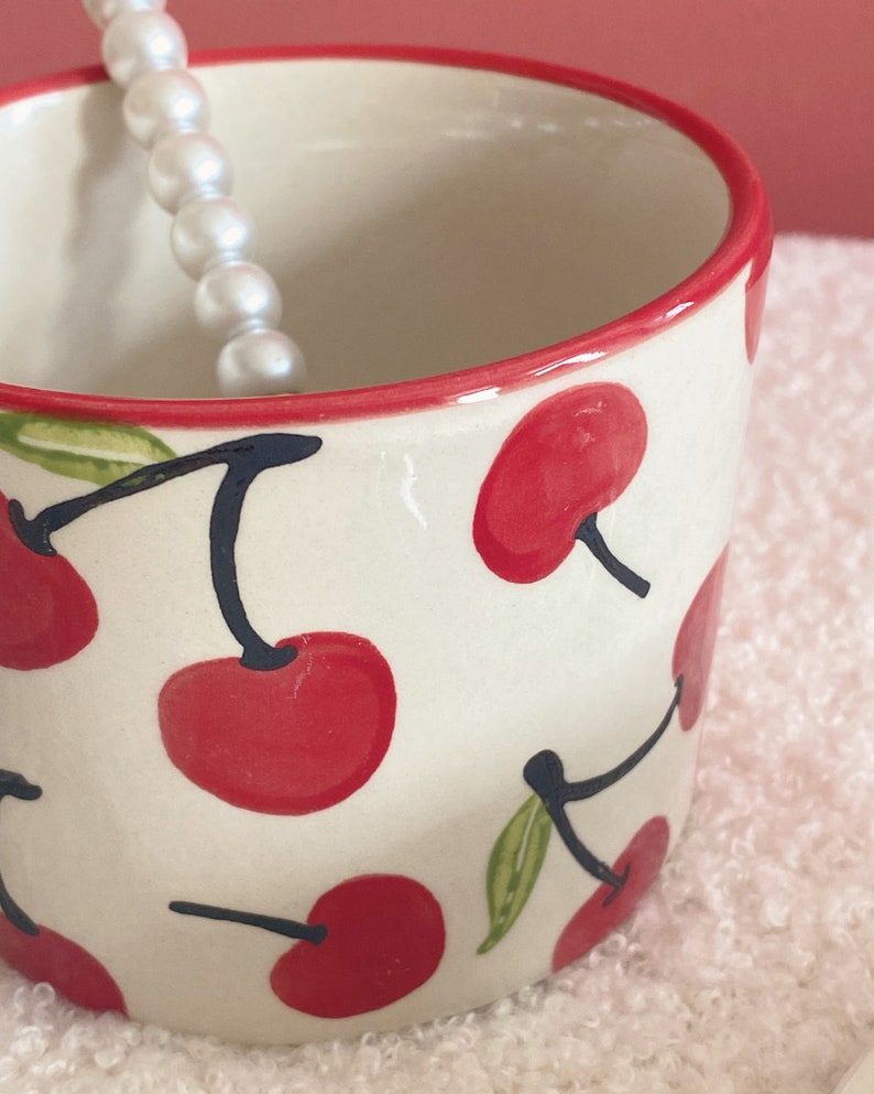 May include: A white ceramic mug with a red rim and a cherry pattern. The mug is decorated with red cherries and green leaves on a white background.