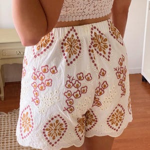 K&ouml;nnte beinhalten: Wei&szlig;e H&auml;kelshorts mit gewellten Kanten und gestickten Blumenmustern in Rosa-, Braun- und Wei&szlig;t&ouml;nen.
