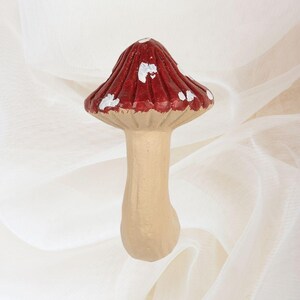 May include: A decorative mushroom figurine with a red cap and white spots. The stem is beige, and the cap has a ribbed texture. The figurine is set against a soft, white background.