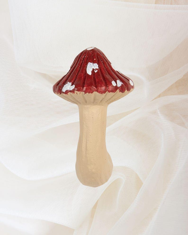 May include: A decorative mushroom figurine with a red cap and white spots. The stem is beige, and the cap has a ribbed texture. The figurine is set against a soft, white background.