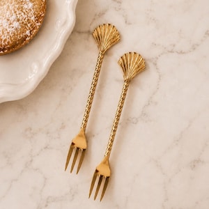 May include: Two gold-toned metal dessert forks featuring a twisted handle design and a shell-shaped finial at the top. The forks have three tines each and a polished finish, presented alongside a white plate with a pastry dusted in sugar.