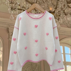 May include: A white sweater with pink embroidered hearts. The sweater has a crew neck and long sleeves.