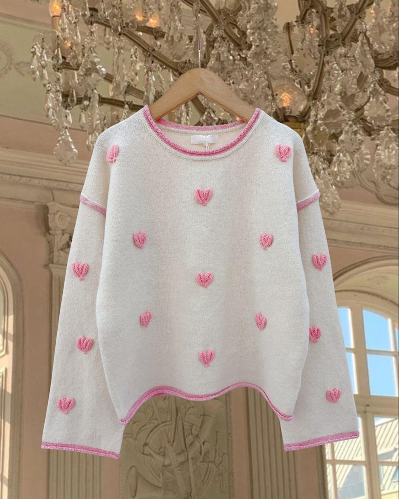 May include: A white sweater with pink embroidered hearts. The sweater has a crew neck and long sleeves.
