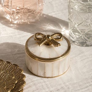 Puede incluir: Un joyero redondo de n&aacute;car blanco con un borde de metal dorado y un asa decorativa en forma de lazo dorado en la tapa. La caja presenta paneles verticales de material de concha iridiscente y un acabado met&aacute;lico pulido en el lazo de la tapa.