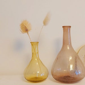 May include: Two glass vases, one amber and one clear, with dried flowers. The amber vase has a round body and a thin neck. The clear vase has a wider base and a long, thin neck. Both vases are sitting on a white surface.