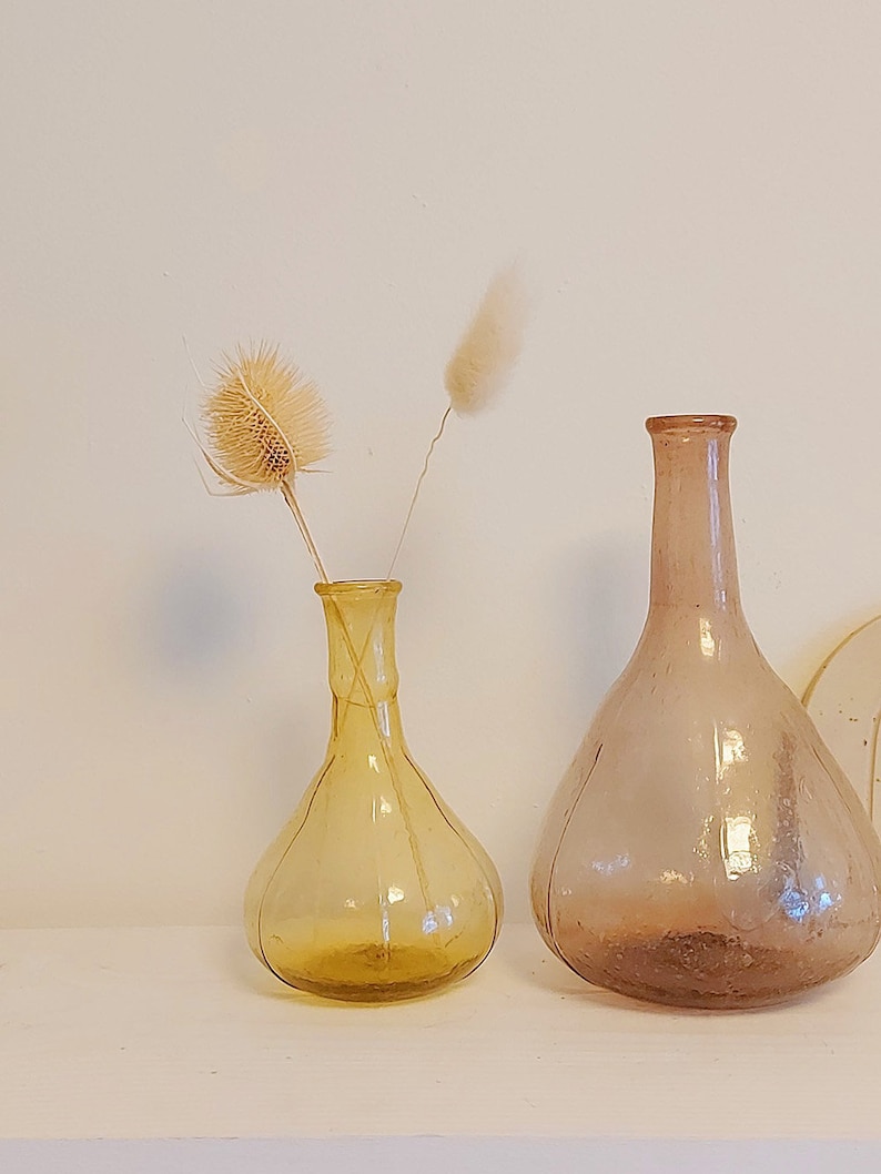 May include: Two glass vases, one amber and one clear, with dried flowers. The amber vase has a round body and a thin neck. The clear vase has a wider base and a long, thin neck. Both vases are sitting on a white surface.