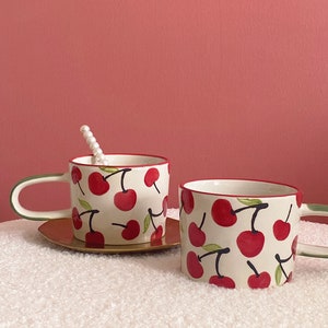 May include: Two white ceramic mugs with a red cherry pattern. The mugs have green handles and a red rim. The mugs are sitting on a white fluffy surface.