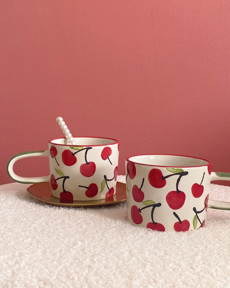 May include: Two white ceramic mugs with a red cherry pattern. The mugs have green handles and a red rim. The mugs are sitting on a white fluffy surface.