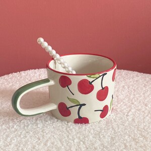May include: A white ceramic mug with a green handle and a red rim. The mug is decorated with a pattern of red cherries with green stems and leaves.