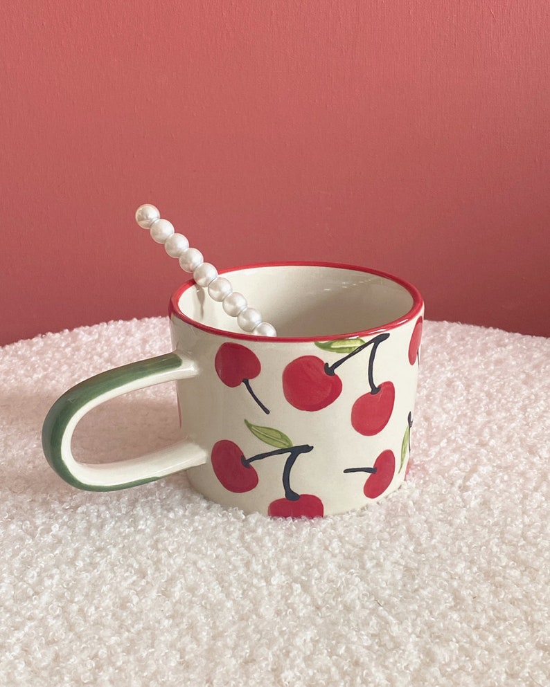 May include: A white ceramic mug with a green handle and a red rim. The mug is decorated with a pattern of red cherries with green stems and leaves.