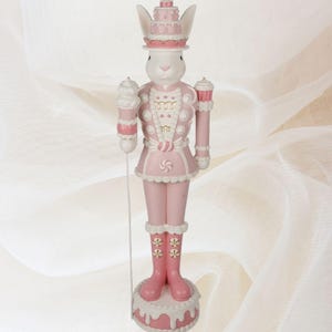 Puede incluir: Un cascanueces de conejito rosa y blanco con un sombrero de pastel y un bastón de caramelo en la mano. El cascanueces está de pie sobre una base de pastel rosa y blanco.