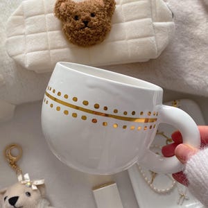 May include: White ceramic mug with gold dot pattern. A plush teddy bear keychain and a white cosmetic case with a teddy bear applique are also visible. The mug is held by a person wearing a white sweater.