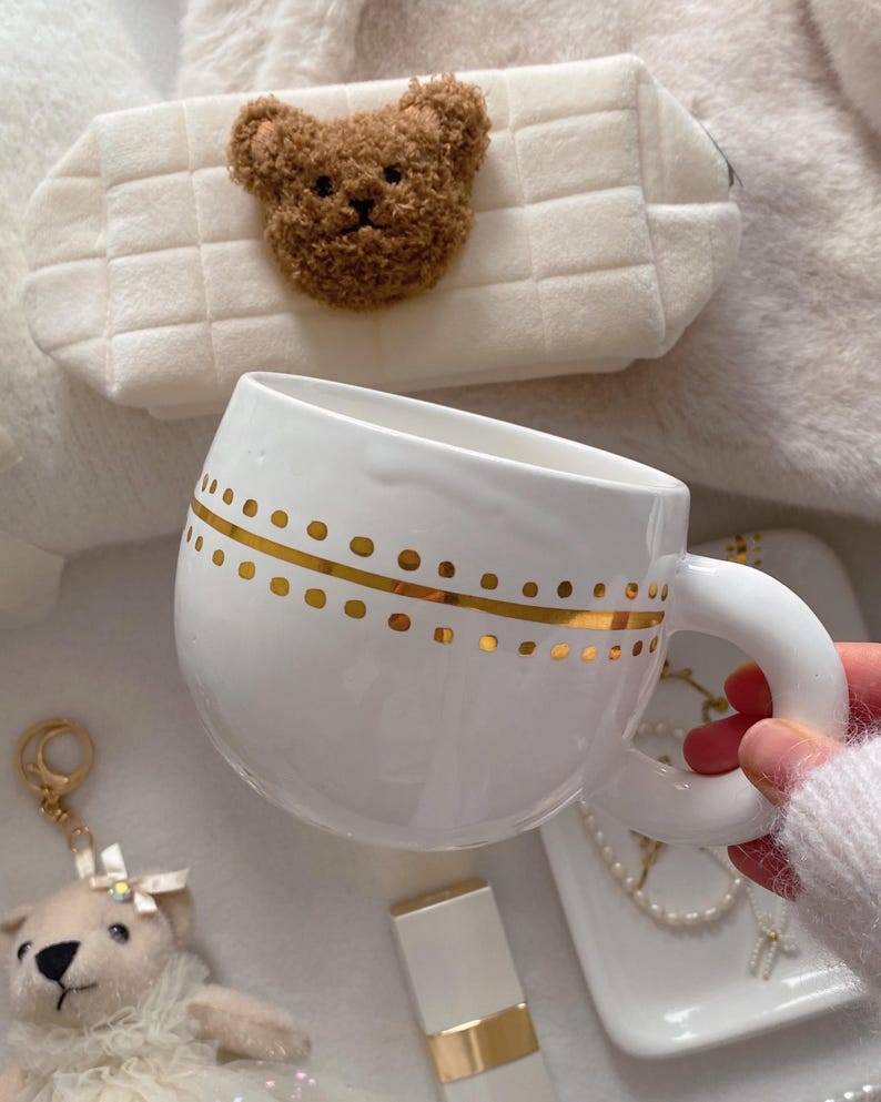 May include: White ceramic mug with gold dot pattern. A plush teddy bear keychain and a white cosmetic case with a teddy bear applique are also visible. The mug is held by a person wearing a white sweater.