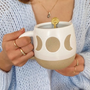 May include: White ceramic mug with a brown speckled glaze and a moon phase design. The mug is being held by a person wearing a light blue knitted cardigan.