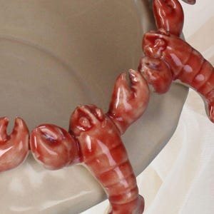 May include: A beige ceramic serving dish with a decorative border of red lobster figures. The lobsters are detailed with claws and segmented bodies, adding a nautical theme. The dish has a smooth, glossy finish.