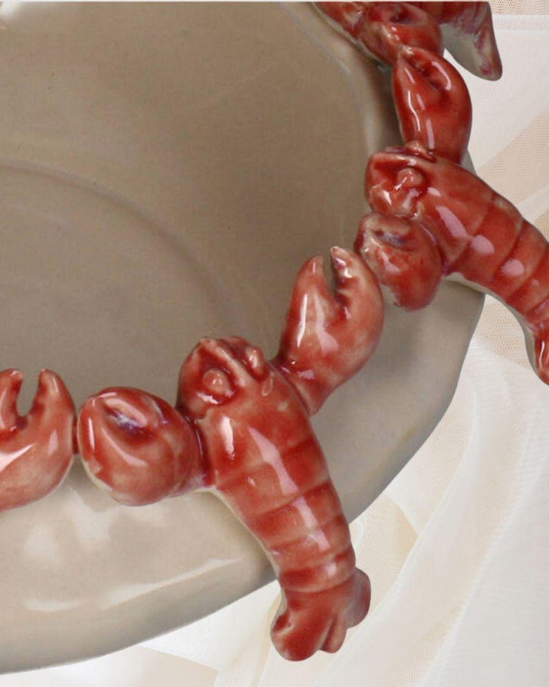 May include: A beige ceramic serving dish with a decorative border of red lobster figures. The lobsters are detailed with claws and segmented bodies, adding a nautical theme. The dish has a smooth, glossy finish.
