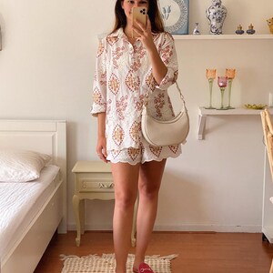 K&ouml;nnte beinhalten: Ein wei&szlig;es, besticktes Hemd- und Shorts-Set mit einer wei&szlig;en, halbmondf&ouml;rmigen Handtasche und pinkfarbenen Leder-Slipper.