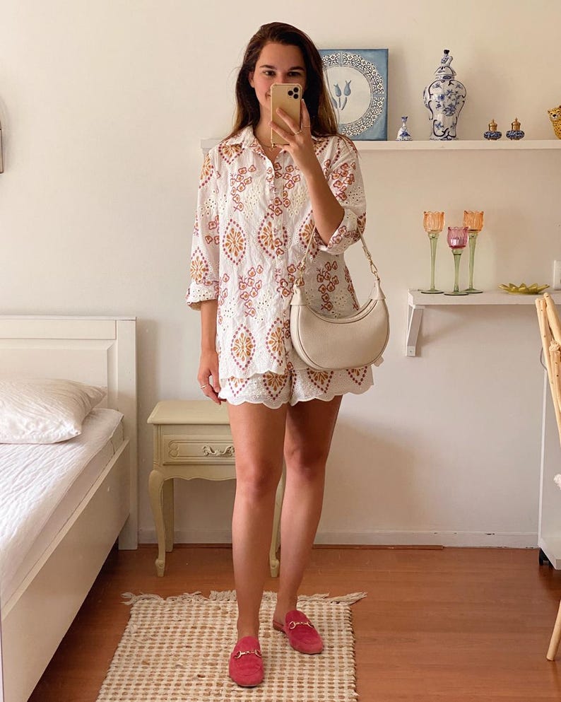 K&ouml;nnte beinhalten: Ein wei&szlig;es, besticktes Hemd- und Shorts-Set mit einer wei&szlig;en, halbmondf&ouml;rmigen Handtasche und pinkfarbenen Leder-Slipper.
