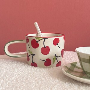 May include: A white ceramic mug with a red rim and a green handle. The mug is decorated with a pattern of red cherries on a white background. A white pearl-like spoon is resting on the rim of the mug.