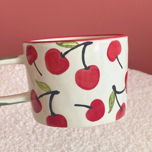 May include: White ceramic mug with a red rim and a pattern of red cherries with green leaves and black stems.