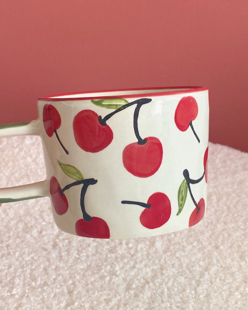 May include: White ceramic mug with a red rim and a pattern of red cherries with green leaves and black stems.