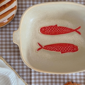 May include: A cream-colored ceramic serving dish with two red fish designs. The dish has a rectangular shape with rounded edges and small handles. The fish are detailed with scales and fins.