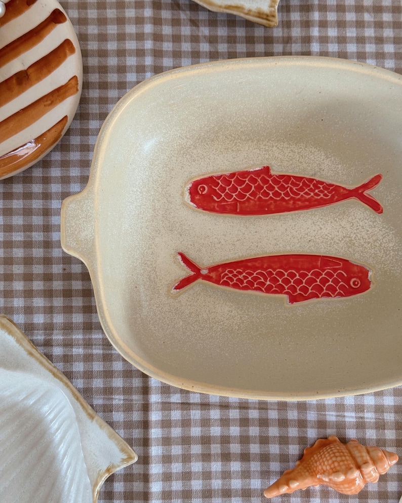 May include: A cream-colored ceramic serving dish with two red fish designs. The dish has a rectangular shape with rounded edges and small handles. The fish are detailed with scales and fins.