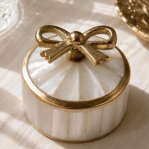 Puede incluir: Un joyero redondo con incrustaciones de n&aacute;car blanco dispuestas en un patr&oacute;n de rayas verticales. El recipiente est&aacute; enmarcado con un borde de metal dorado y rematado con un asa decorativa en forma de lazo de metal dorado en la tapa.