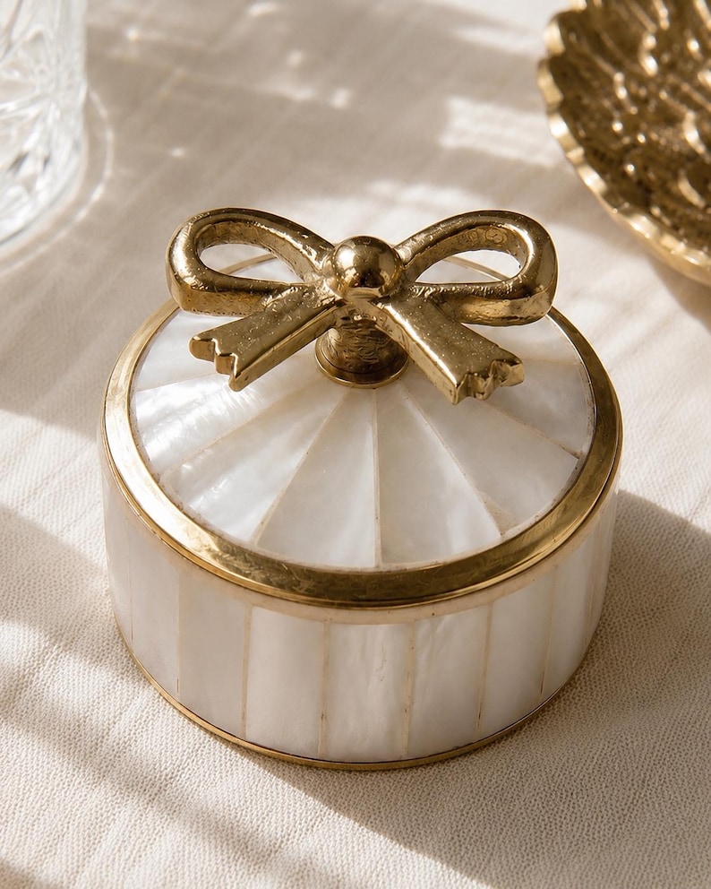 Puede incluir: Un joyero redondo con incrustaciones de n&aacute;car blanco dispuestas en un patr&oacute;n de rayas verticales. El recipiente est&aacute; enmarcado con un borde de metal dorado y rematado con un asa decorativa en forma de lazo de metal dorado en la tapa.