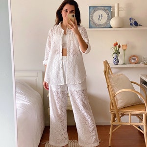 Puede incluir: Un conjunto de top y pantalón blanco de encaje con un ajuste holgado. El top tiene cuello y mangas largas. El pantalón es de pierna ancha y tiene una pierna recta.