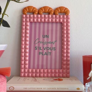 Puede incluir: Marco de fotos rosa con un diseño texturizado en relieve y un fondo a rayas blanco y rosa. El marco tiene una fila de adornos en forma de croissant naranja en la parte superior. El texto dentro del marco dice "Un Croissant S'il Vous Plait".
