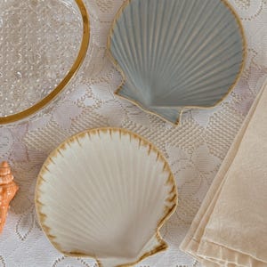 May include: Two ceramic seashell-shaped dishes. One is light blue, the other is off-white. Both have a textured, ridged design and a brown rim. The dishes are on a white lace tablecloth, with a clear glass dish with a gold rim in the background.