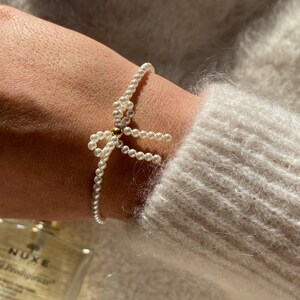 May include: Delicate pearl bracelet featuring a bow design. The bracelet is made of small, white pearls and has a gold-colored accent in the center of the bow. The bracelet is worn on a wrist, and the background is a soft, neutral color.