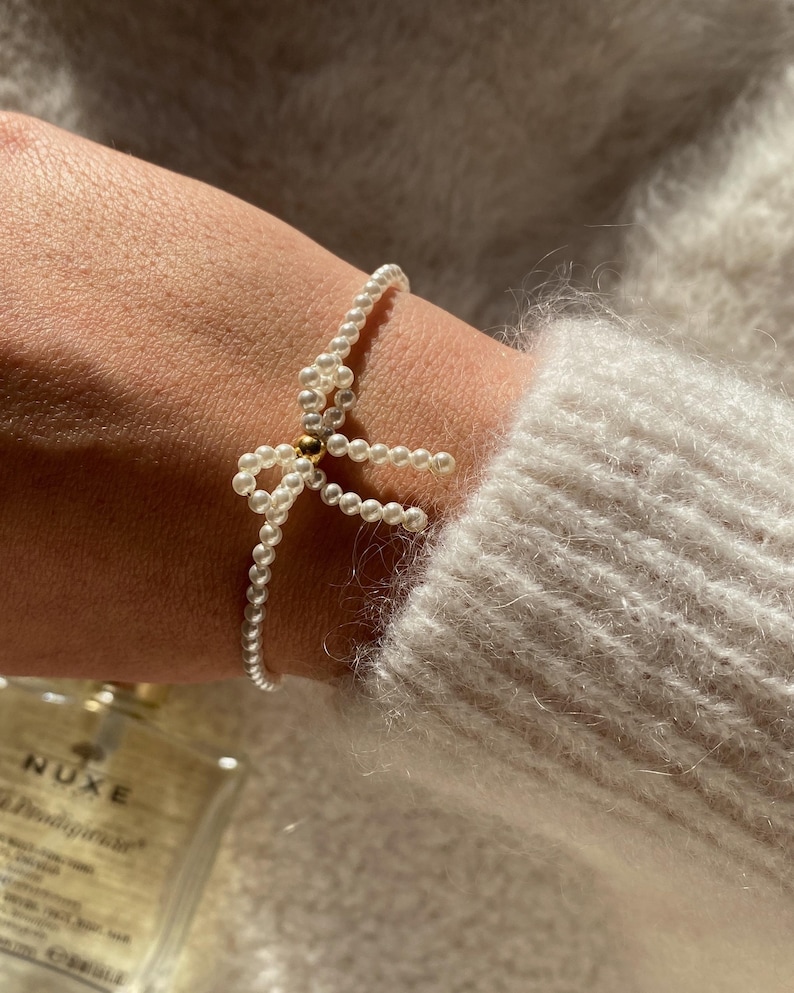 May include: Delicate pearl bracelet featuring a bow design. The bracelet is made of small, white pearls and has a gold-colored accent in the center of the bow. The bracelet is worn on a wrist, and the background is a soft, neutral color.