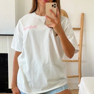 K&ouml;nnte beinhalten: Ein wei&szlig;es Oversize-T-Shirt mit einem rosafarbenen "California"-Aufdruck auf der Vorderseite. Das Model tr&auml;gt das Shirt zu Jeans-Shorts.