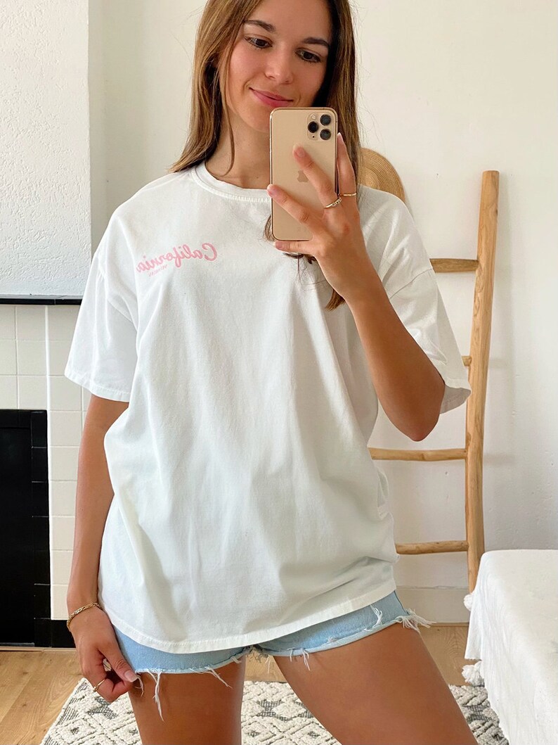 K&ouml;nnte beinhalten: Ein wei&szlig;es Oversize-T-Shirt mit einem rosafarbenen "California"-Aufdruck auf der Vorderseite. Das Model tr&auml;gt das Shirt zu Jeans-Shorts.
