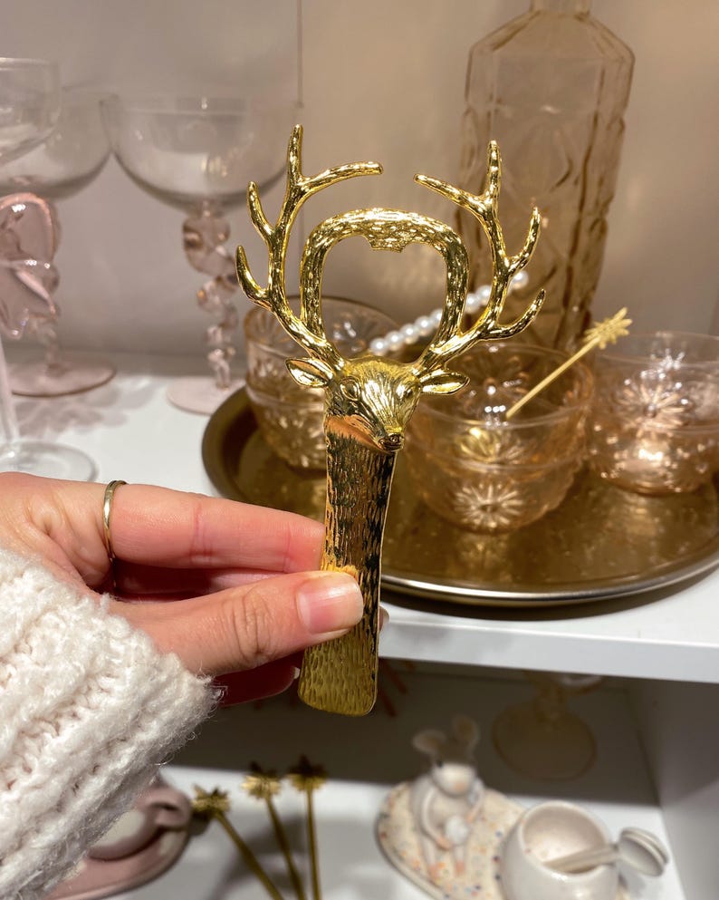 May include: A gold-colored bottle opener with a deer head design. The opener features detailed antlers and a textured handle. The background includes glassware and decorative items, suggesting a bar or home decor theme.