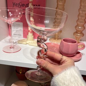 May include: A clear champagne glass with a twisted stem is held in front of a shelf with other pink glassware. The glass has a wide bowl and a clear, spiral-shaped stem. Other items include a pink teacup and saucer, and a glass with white dots.