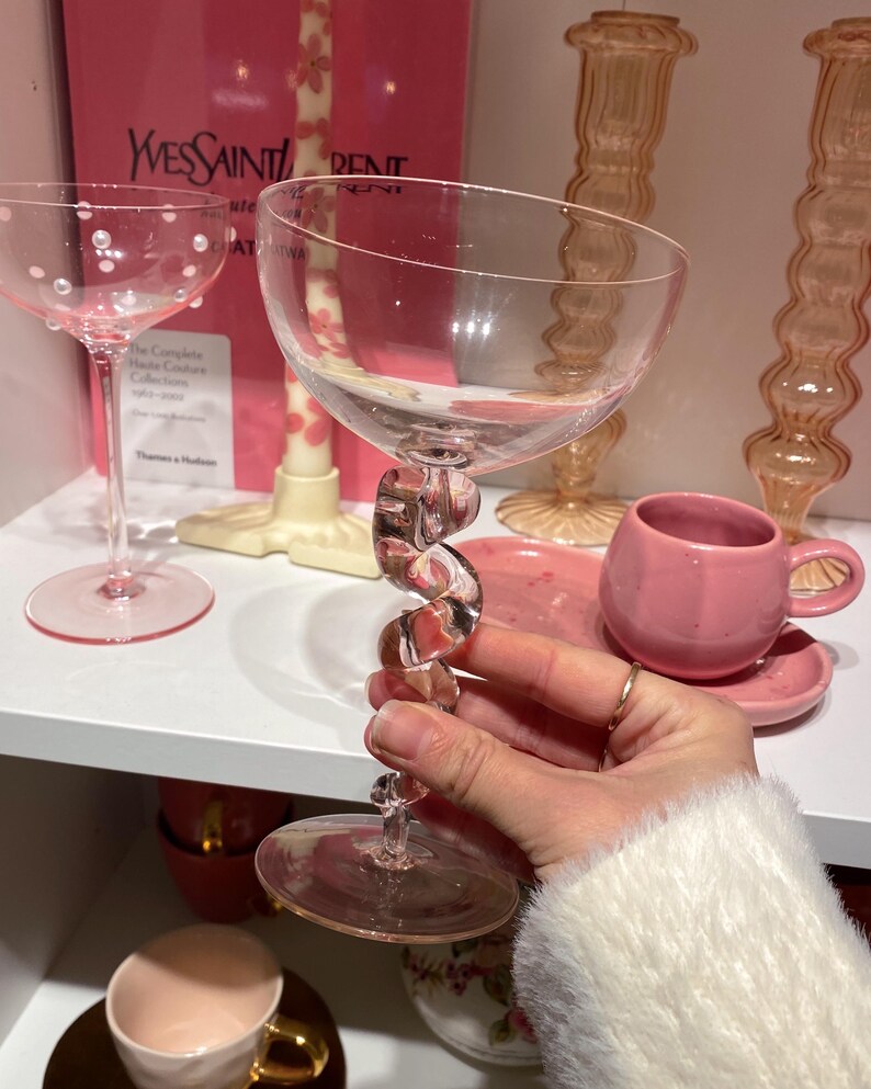 May include: A clear champagne glass with a twisted stem is held in front of a shelf with other pink glassware. The glass has a wide bowl and a clear, spiral-shaped stem. Other items include a pink teacup and saucer, and a glass with white dots.