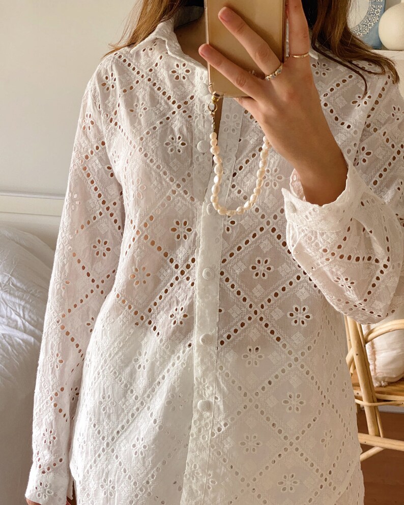 May include: White long-sleeved button-up shirt with a delicate eyelet pattern. The shirt has a relaxed fit and a collar.