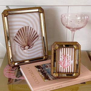 Puede incluir: Dos cuadros dorados con ilustraciones de conchas marinas y corales, apoyados en una pila de libros. También se ve una copa de champán rosa con lunares. Los marcos tienen un borde espejado.