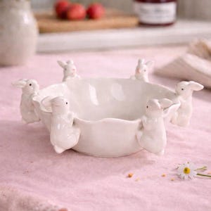 Puede incluir: Un cuenco de cerámica blanco con borde festoneado, adornado con seis pequeñas figuras de conejitos blancos. El cuenco está sobre una superficie rosa, con un fondo borroso que muestra flores y un tarro de mermelada. Una encantadora pieza decorativa con temática de Pascua.