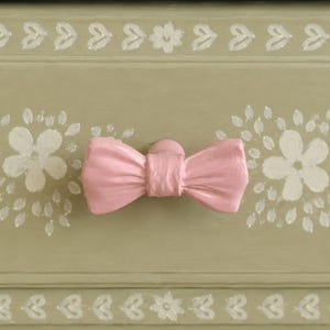 May include: A sage green drawer front featuring a hand-painted white floral pattern and a horizontal border of small white hearts. A solid light pink bow-shaped knob is centered on the drawer, serving as the functional handle for the furniture piece.