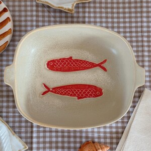 May include: A cream-colored ceramic serving dish with two red fish designs. The dish has a rectangular shape with rounded corners and handles. The background includes a checkered tablecloth and seashell decorations.