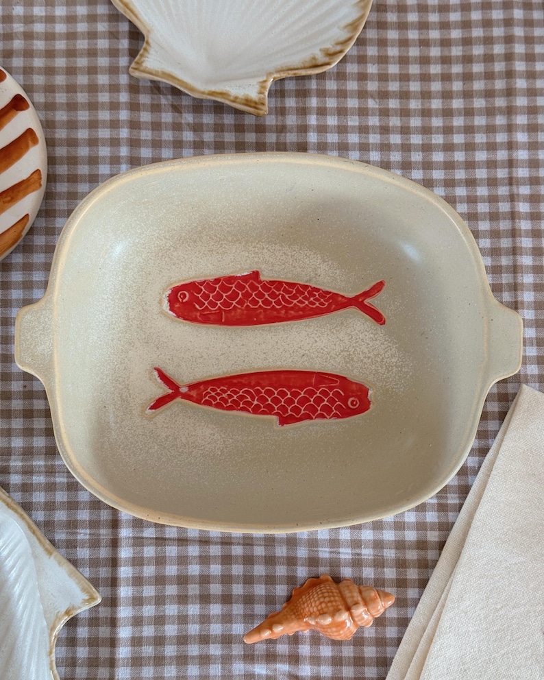May include: A cream-colored ceramic serving dish with two red fish designs. The dish has a rectangular shape with rounded corners and handles. The background includes a checkered tablecloth and seashell decorations.