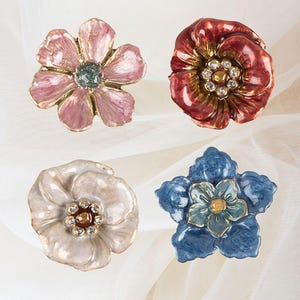 Peut inclure: Quatre broches en forme de fleurs en émail avec des centres ornés de bijoux. Les broches sont dans des tons de rose, rouge, blanc et bleu.