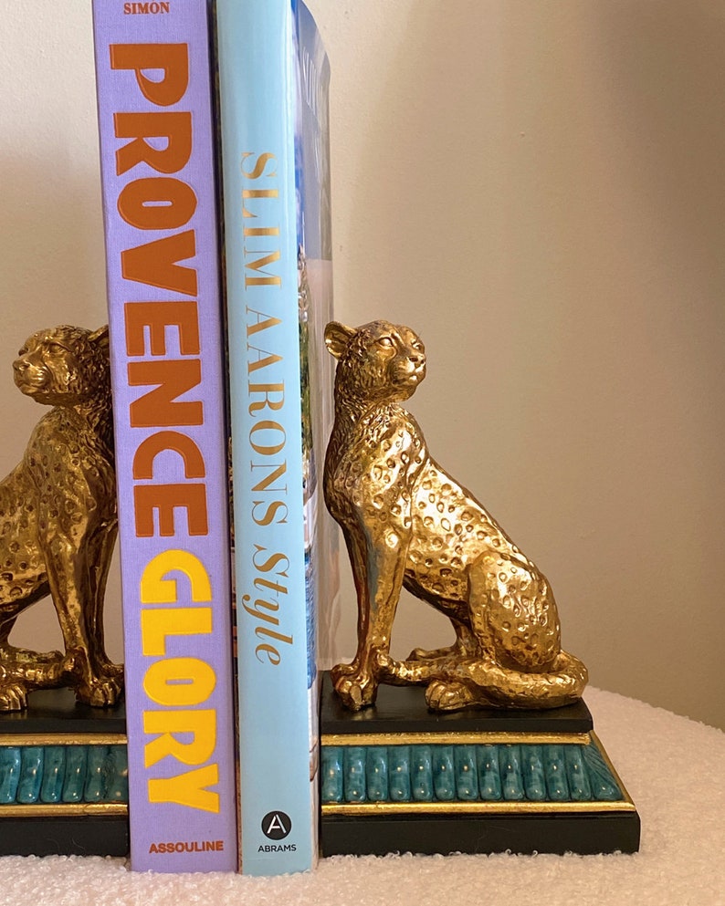 K&ouml;nnte beinhalten: Goldfarbene Geparden-Buchst&uuml;tzen mit schwarzem und t&uuml;rkisem Sockel, die zwei B&uuml;cher halten. Die B&uuml;cher tragen die Titel "Provence Glory" und "Slim Aarons Style". Die Geparden sitzen und sind dekorativ.