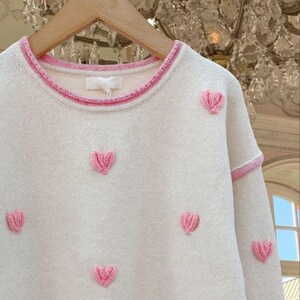 May include: A cream-colored sweater with pink heart-shaped embroidery. The sweater has a pink trim around the neckline.