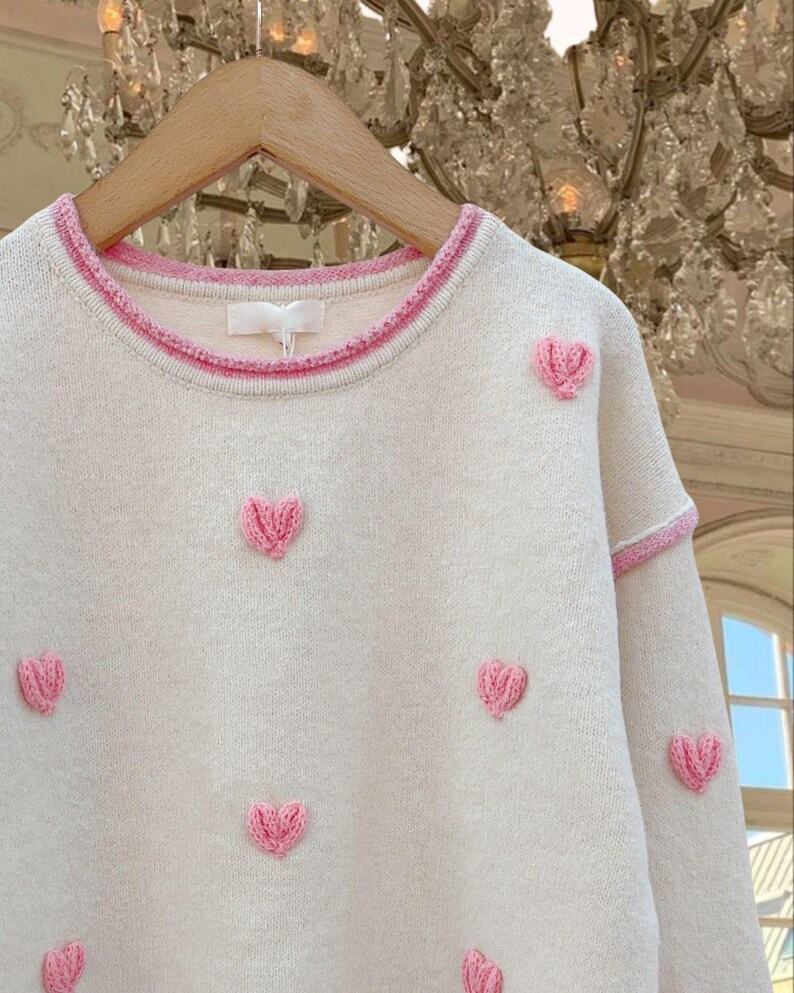 May include: A cream-colored sweater with pink heart-shaped embroidery. The sweater has a pink trim around the neckline.
