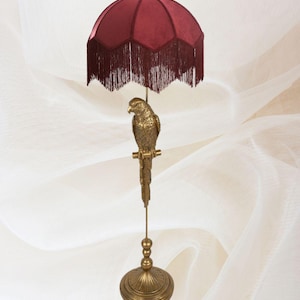 Puede incluir: Una lámpara decorativa con un loro dorado posado sobre un soporte. La lámpara tiene una pantalla de color burdeos con flecos. La base y el soporte también son dorados, creando una estética vintage.
